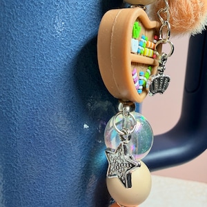 K&ouml;nnte beinhalten: Ein Charm mit einem Miniatur-B&uuml;cherregal, einer silbernen Krone und einem Sternanh&auml;nger. Der Charm ist an einer Perlenkette mit pfirsichfarbenen, cremefarbenen und irisierenden Perlen befestigt. Ein flauschiger orangefarbener Pompon und ein silberner Charm mit der Aufschrift "bride" sind ebenfalls enthalten.