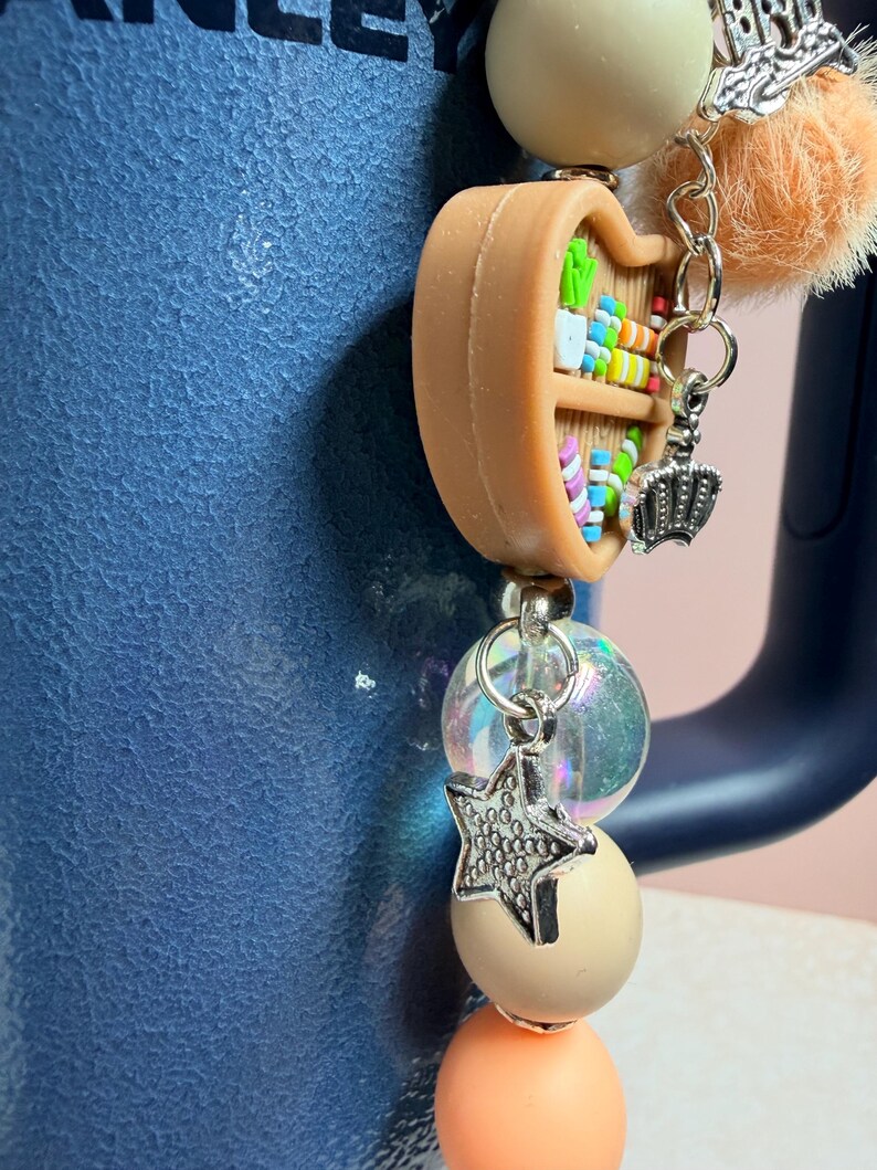 K&ouml;nnte beinhalten: Ein Charm mit einem Miniatur-B&uuml;cherregal, einer silbernen Krone und einem Sternanh&auml;nger. Der Charm ist an einer Perlenkette mit pfirsichfarbenen, cremefarbenen und irisierenden Perlen befestigt. Ein flauschiger orangefarbener Pompon und ein silberner Charm mit der Aufschrift "bride" sind ebenfalls enthalten.