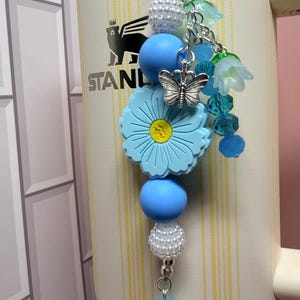May include: A decorative tumbler charm featuring a large light blue flower bead, smooth light blue round beads, white textured beads, and translucent blue flower-shaped beads. A silver-toned butterfly charm hangs from the center of the arrangement.