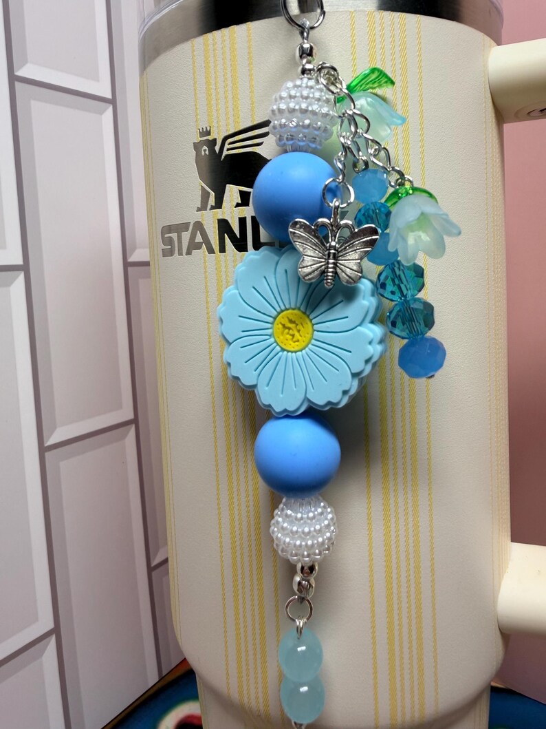 May include: A decorative tumbler charm featuring a large light blue flower bead, smooth light blue round beads, white textured beads, and translucent blue flower-shaped beads. A silver-toned butterfly charm hangs from the center of the arrangement.