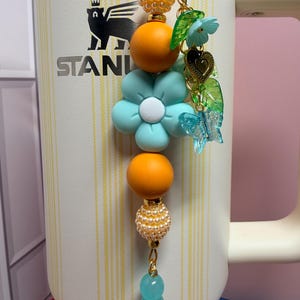 May include: A decorative tumbler charm featuring a vertical arrangement of orange, light blue, and pearl-textured beads, accented with a light blue flower, a gold heart, green leaves, and a clear butterfly charm, attached to a white Stanley tumbler.