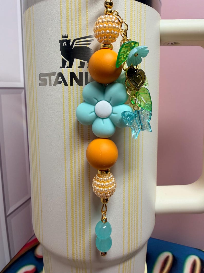 May include: A decorative tumbler charm featuring a vertical arrangement of orange, light blue, and pearl-textured beads, accented with a light blue flower, a gold heart, green leaves, and a clear butterfly charm, attached to a white Stanley tumbler.
