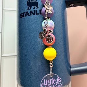 May include: A blue insulated tumbler with a beaded charm. The charm features purple, pink, yellow, and plaid beads, a butterfly charm, and a bicycle charm. A clear acrylic disc with the words "Hello Spring" in purple is attached.