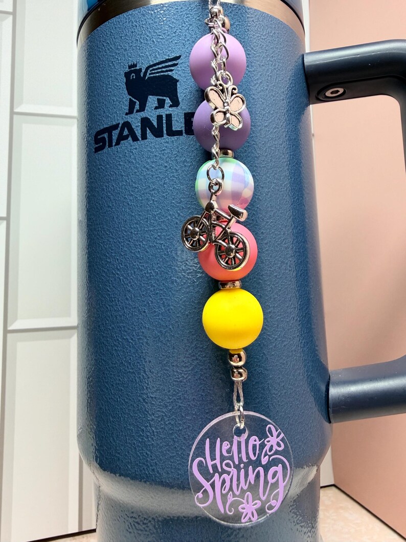 May include: A blue insulated tumbler with a beaded charm. The charm features purple, pink, yellow, and plaid beads, a butterfly charm, and a bicycle charm. A clear acrylic disc with the words "Hello Spring" in purple is attached.