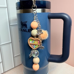 K&ouml;nnte beinhalten: Ein blauer Stanley-Becher mit einem silbernen Anh&auml;nger. Der Anh&auml;nger hat pfirsich- und cremefarbene Perlen, ein herzf&ouml;rmiges B&uuml;cherregal, eine Krone und einen kleinen orangefarbenen Pompon. Der Becher hat einen Griff und einen Strohhalm.