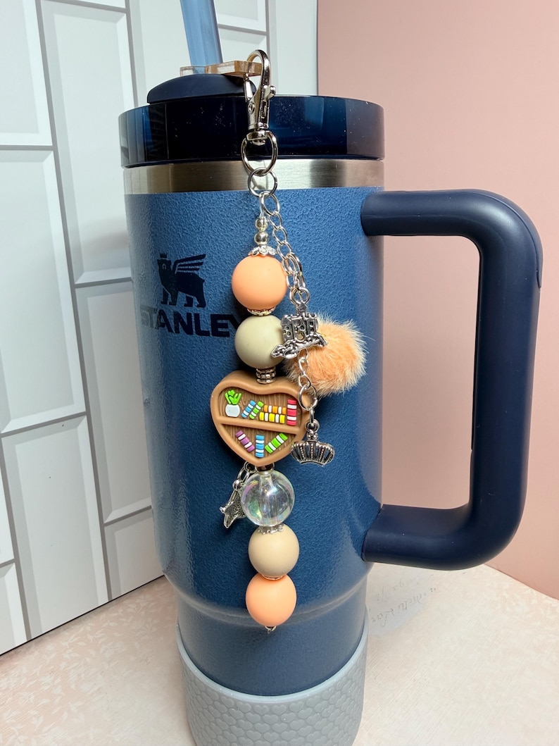 K&ouml;nnte beinhalten: Ein blauer Stanley-Becher mit einem silbernen Anh&auml;nger. Der Anh&auml;nger hat pfirsich- und cremefarbene Perlen, ein herzf&ouml;rmiges B&uuml;cherregal, eine Krone und einen kleinen orangefarbenen Pompon. Der Becher hat einen Griff und einen Strohhalm.