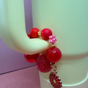 Op de afbeelding: Een rode kralenarmband met gouden accenten en een bloembedel. De armband heeft grote, ronde, glanzende rode kralen, een getextureerde rode kraal en een kleine roze bloemkraal. Een rode en gouden bloembedel bungelt aan de armband.