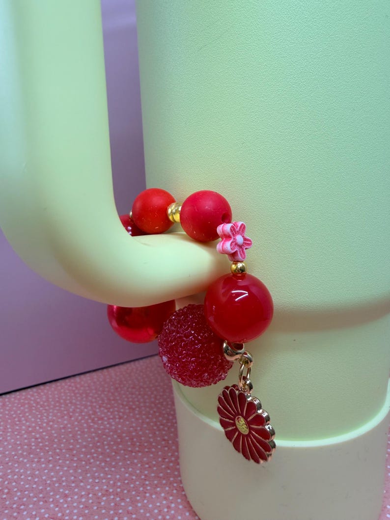 Op de afbeelding: Een rode kralenarmband met gouden accenten en een bloembedel. De armband heeft grote, ronde, glanzende rode kralen, een getextureerde rode kraal en een kleine roze bloemkraal. Een rode en gouden bloembedel bungelt aan de armband.