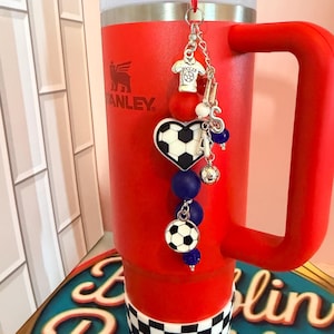 Puede incluir: Un vaso t&eacute;rmico de color rojo brillante con asa y tapa blanca, que presenta una base a cuadros blancos y negros. Un dije decorativo cuelga de la tapa, mostrando un dije de camiseta de f&uacute;tbol, un bal&oacute;n en forma de coraz&oacute;n y cuentas azules.