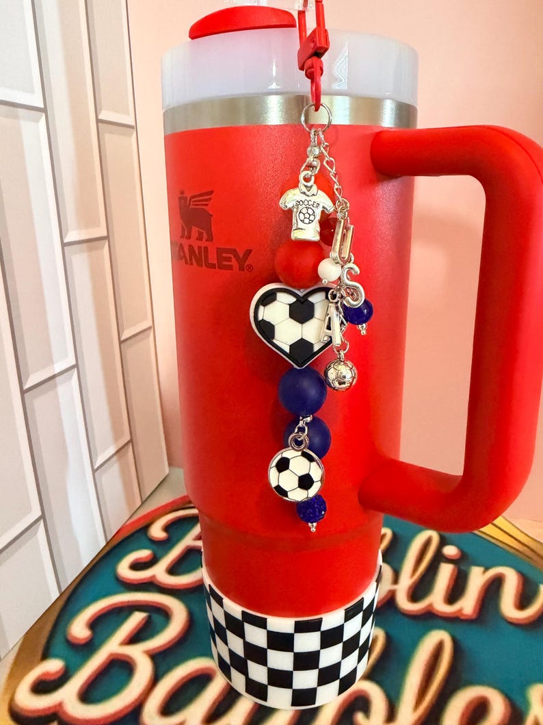 Puede incluir: Un vaso t&eacute;rmico de color rojo brillante con asa y tapa blanca, que presenta una base a cuadros blancos y negros. Un dije decorativo cuelga de la tapa, mostrando un dije de camiseta de f&uacute;tbol, un bal&oacute;n en forma de coraz&oacute;n y cuentas azules.