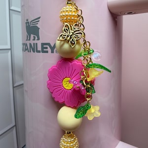 May include: A decorative tumbler charm featuring a gold-tone chain with two textured gold beads, two smooth cream-colored beads, a bright pink flower-shaped silicone bead, a gold butterfly charm, and small translucent green and yellow flower beads.