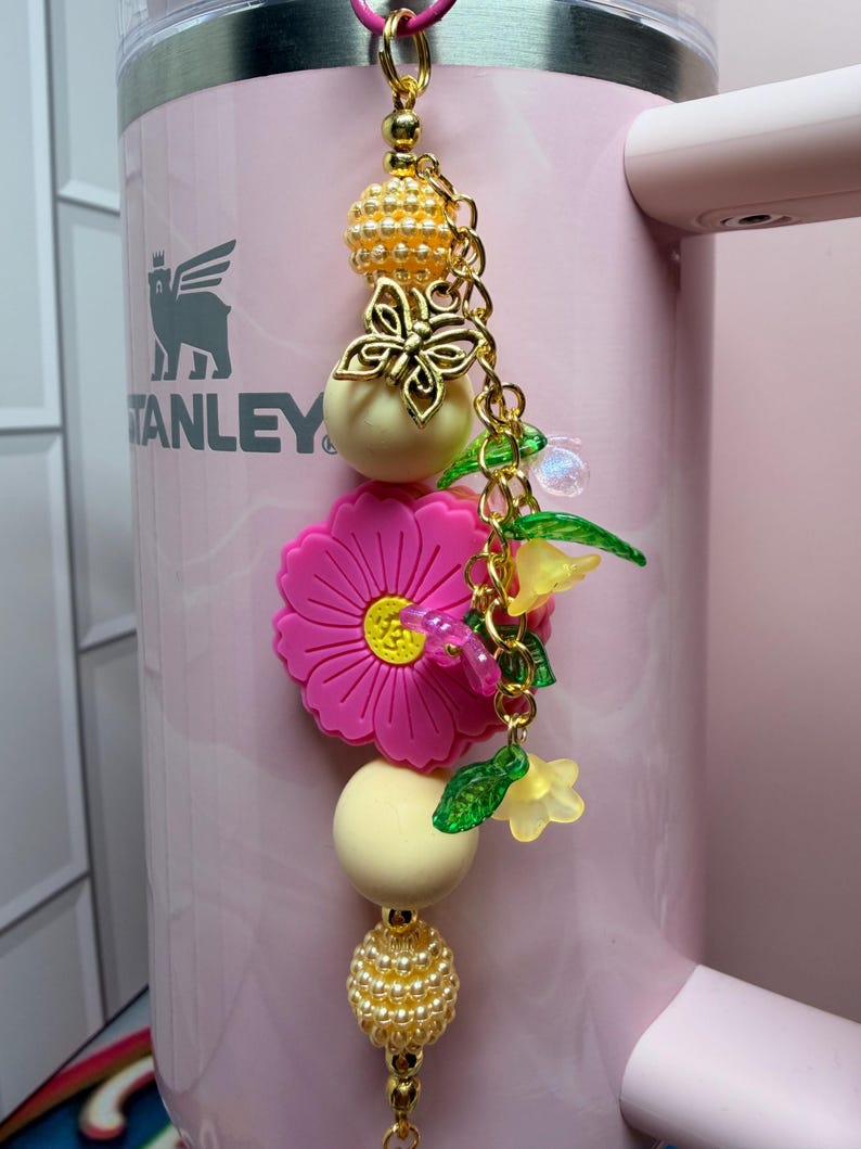 May include: A decorative tumbler charm featuring a gold-tone chain with two textured gold beads, two smooth cream-colored beads, a bright pink flower-shaped silicone bead, a gold butterfly charm, and small translucent green and yellow flower beads.