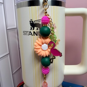 May include: A decorative beaded charm hanging from a cream-colored Stanley tumbler. The charm features a pink flamingo pendant, a peach-colored flower bead, and alternating dark green and bright pink textured beads on a gold-toned metal chain.
