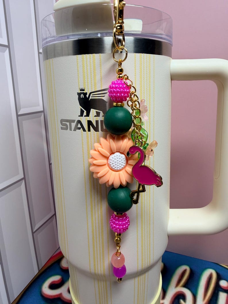 May include: A decorative beaded charm hanging from a cream-colored Stanley tumbler. The charm features a pink flamingo pendant, a peach-colored flower bead, and alternating dark green and bright pink textured beads on a gold-toned metal chain.