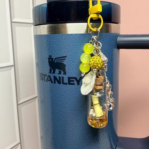 May include: A dark blue Stanley tumbler with a yellow clip-on charm featuring three yellow beads, a yellow glitter sphere, a small iridescent shell, a silver seahorse charm, and a miniature glass bottle containing a yellow scroll and gold glitter.