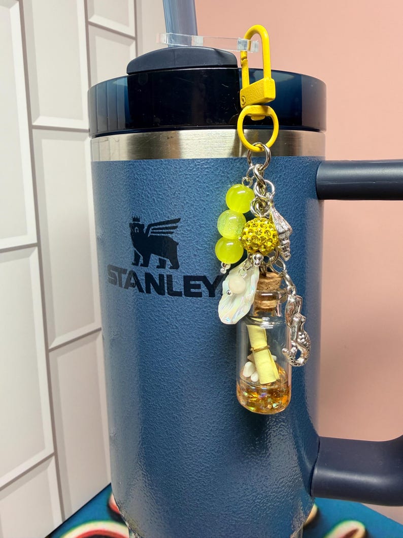May include: A dark blue Stanley tumbler with a yellow clip-on charm featuring three yellow beads, a yellow glitter sphere, a small iridescent shell, a silver seahorse charm, and a miniature glass bottle containing a yellow scroll and gold glitter.
