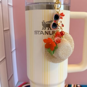 May include: A cream Stanley tumbler with a yellow striped pattern, adorned with a gold keychain charm. The charm features miniature sunglasses, flowers, and a small straw hat. The tumbler has a white handle and a clear lid.