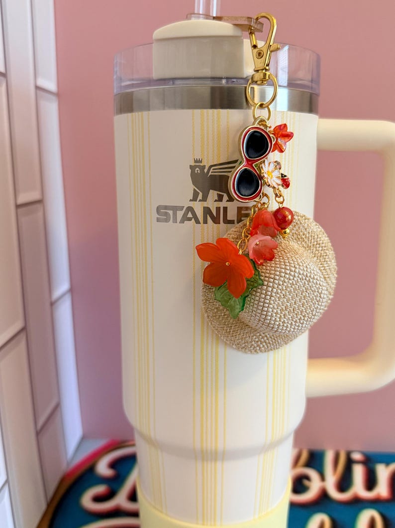 May include: A cream Stanley tumbler with a yellow striped pattern, adorned with a gold keychain charm. The charm features miniature sunglasses, flowers, and a small straw hat. The tumbler has a white handle and a clear lid.