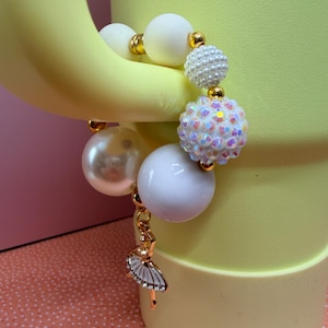 Op de afbeelding: Een kralenarmband met grote witte en parelmoeren kralen, geaccentueerd met goudkleurige afstandhouders. Een bedel van een ballerina in een tutu hangt aan de armband. De armband wordt gepresenteerd tegen een gele achtergrond.