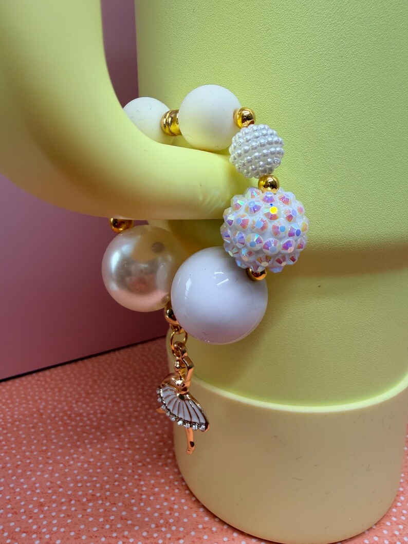 Op de afbeelding: Een kralenarmband met grote witte en parelmoeren kralen, geaccentueerd met goudkleurige afstandhouders. Een bedel van een ballerina in een tutu hangt aan de armband. De armband wordt gepresenteerd tegen een gele achtergrond.