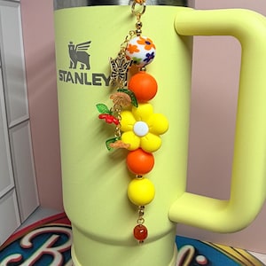 May include: A yellow Stanley tumbler with a handle and a decorative beaded charm attached to the lid. The charm features a gold-tone butterfly, a large yellow flower, orange and yellow round beads, and a small white bead with floral patterns.