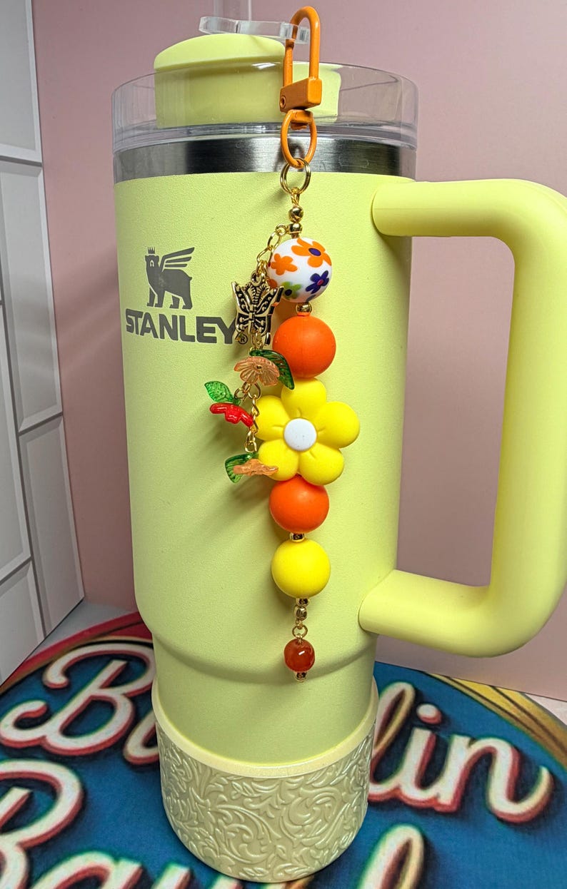 May include: A yellow Stanley tumbler with a handle and a decorative beaded charm attached to the lid. The charm features a gold-tone butterfly, a large yellow flower, orange and yellow round beads, and a small white bead with floral patterns.