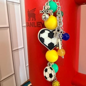 Puede incluir: Un dije para vaso con tem&aacute;tica de f&uacute;tbol que incluye una cadena plateada con cuentas de colores, un dije de coraz&oacute;n con un bal&oacute;n de f&uacute;tbol, un bal&oacute;n redondo y un dije plateado con el texto I LOVE SOCCER, colgado de un vaso rojo brillante.