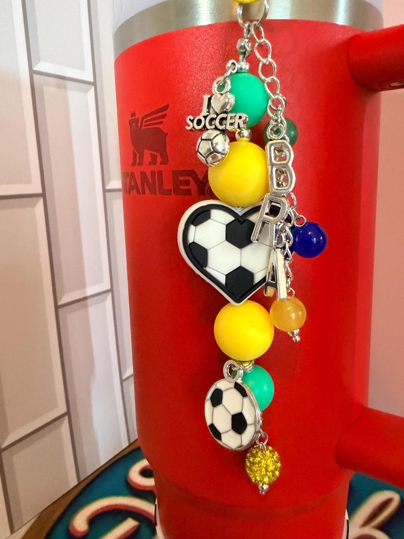 Puede incluir: Un dije para vaso con tem&aacute;tica de f&uacute;tbol que incluye una cadena plateada con cuentas de colores, un dije de coraz&oacute;n con un bal&oacute;n de f&uacute;tbol, un bal&oacute;n redondo y un dije plateado con el texto I LOVE SOCCER, colgado de un vaso rojo brillante.