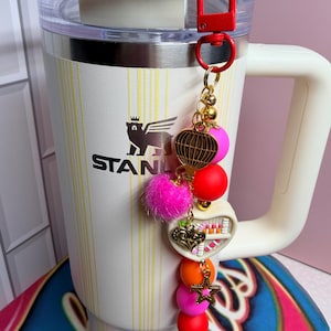 K&ouml;nnte beinhalten: Ein wei&szlig;er Stanley-Becher mit einem dekorativen Anh&auml;nger. Der Anh&auml;nger enth&auml;lt einen roten Verschluss, goldene Perlen, einen rosa Pompon und verschiedene bunte Perlen und Charms, darunter ein Herz und ein Stern.