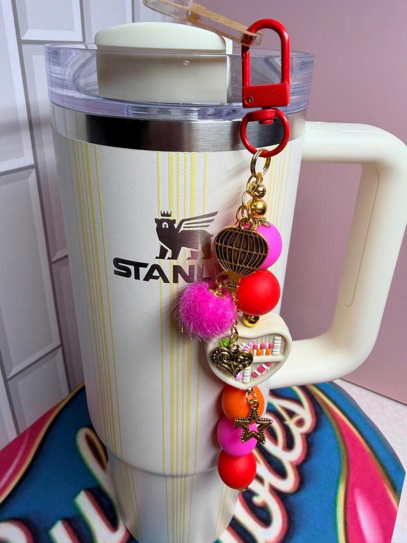 K&ouml;nnte beinhalten: Ein wei&szlig;er Stanley-Becher mit einem dekorativen Anh&auml;nger. Der Anh&auml;nger enth&auml;lt einen roten Verschluss, goldene Perlen, einen rosa Pompon und verschiedene bunte Perlen und Charms, darunter ein Herz und ein Stern.
