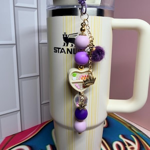 K&ouml;nnte beinhalten: Ein cremefarbener Stanley-Becher mit einem lila-goldenen Perlenanh&auml;nger. Der Anh&auml;nger hat lila und lavendelfarbene Perlen, einen herzf&ouml;rmigen Anh&auml;nger mit einer Krone und einen kleinen lila Pompon. Der Becher hat einen wei&szlig;en Griff und einen Deckel.