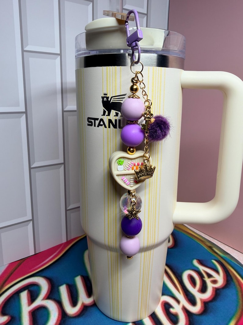 K&ouml;nnte beinhalten: Ein cremefarbener Stanley-Becher mit einem lila-goldenen Perlenanh&auml;nger. Der Anh&auml;nger hat lila und lavendelfarbene Perlen, einen herzf&ouml;rmigen Anh&auml;nger mit einer Krone und einen kleinen lila Pompon. Der Becher hat einen wei&szlig;en Griff und einen Deckel.