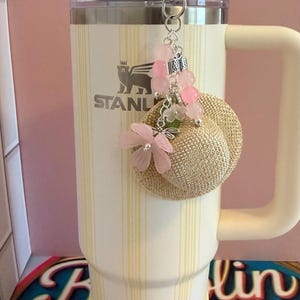 May include: A cream-coloured insulated tumbler with a handle and a straw, decorated with a charm featuring a miniature straw hat, pink beads, and flowers. The tumbler has vertical yellow stripes and the Stanley logo.