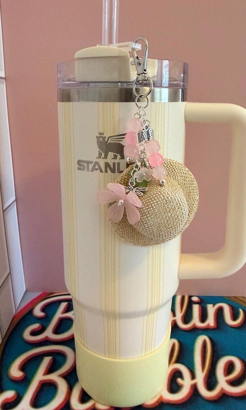 May include: A cream-coloured insulated tumbler with a handle and a straw, decorated with a charm featuring a miniature straw hat, pink beads, and flowers. The tumbler has vertical yellow stripes and the Stanley logo.