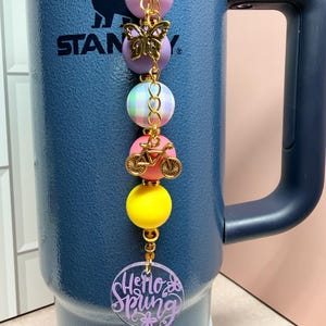 May include: A beaded charm with a gold chain, featuring a butterfly, bicycle, and colourful beads in pink, purple, yellow, and a pastel plaid. A clear, round charm at the bottom reads "Hello Spring" in purple script. The charm is attached to a blue insulated tumbler.
