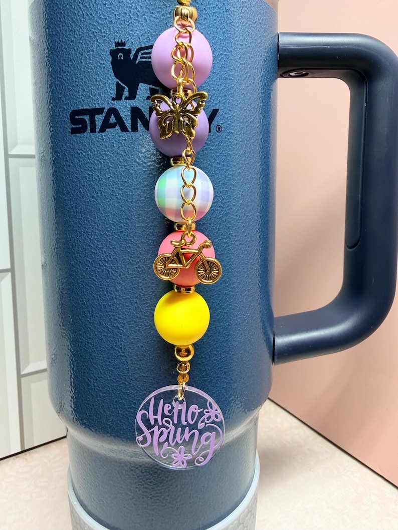 May include: A beaded charm with a gold chain, featuring a butterfly, bicycle, and colourful beads in pink, purple, yellow, and a pastel plaid. A clear, round charm at the bottom reads "Hello Spring" in purple script. The charm is attached to a blue insulated tumbler.