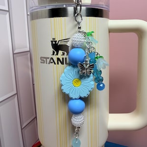 May include: A decorative tumbler charm featuring a silver metal lobster clasp, white pearl-like beads, smooth light blue silicone spheres, a large light blue flower-shaped bead with a yellow center, a silver butterfly charm, and blue floral accents.