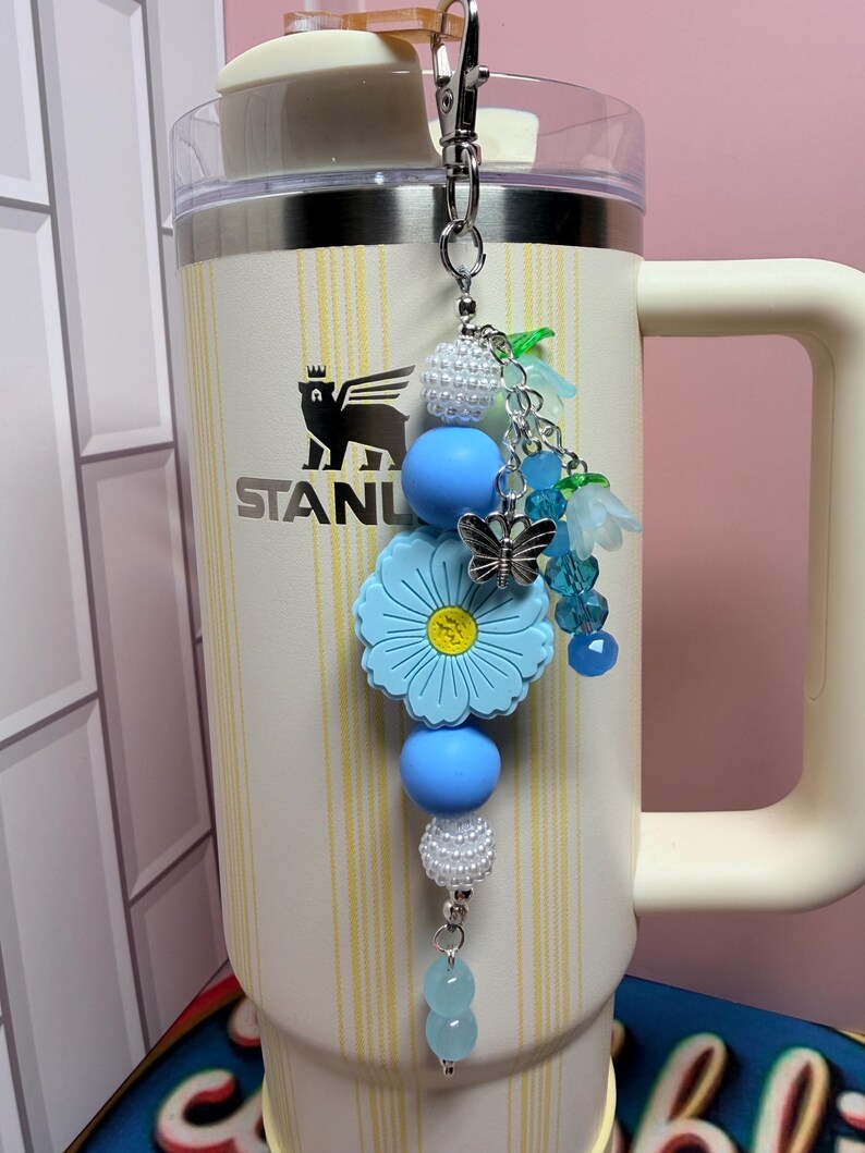 May include: A decorative tumbler charm featuring a silver metal lobster clasp, white pearl-like beads, smooth light blue silicone spheres, a large light blue flower-shaped bead with a yellow center, a silver butterfly charm, and blue floral accents.