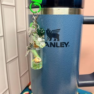 May include: A blue Stanley tumbler with a lime green plastic clip keychain attached. The keychain features silver-toned charms including a sea turtle, a seashell, and a small glass bottle containing a rolled green paper scroll and a tiny seashell.