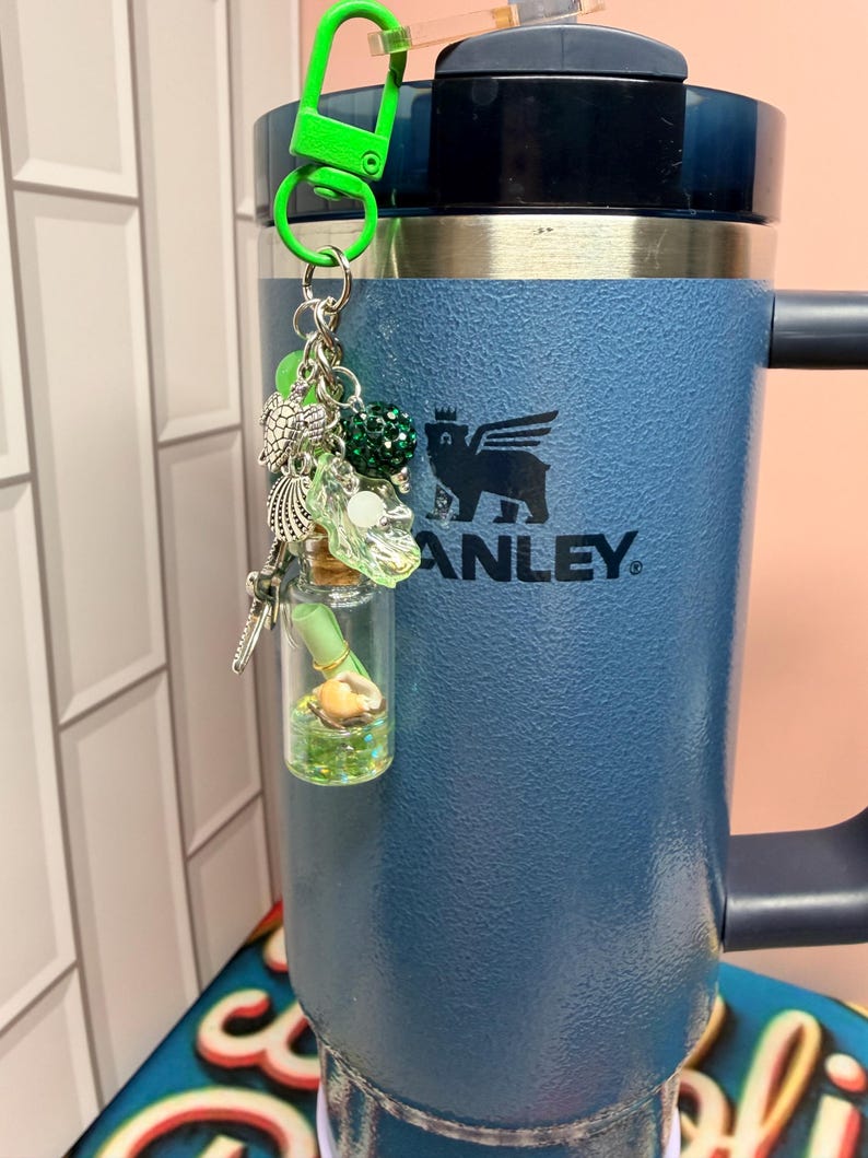 May include: A blue Stanley tumbler with a lime green plastic clip keychain attached. The keychain features silver-toned charms including a sea turtle, a seashell, and a small glass bottle containing a rolled green paper scroll and a tiny seashell.