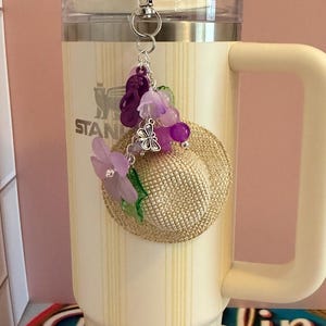 May include: A cream-coloured Stanley tumbler with a straw and handle, decorated with a charm. The charm includes a miniature straw hat, purple flowers, beads, and a butterfly. The tumbler has vertical stripes.