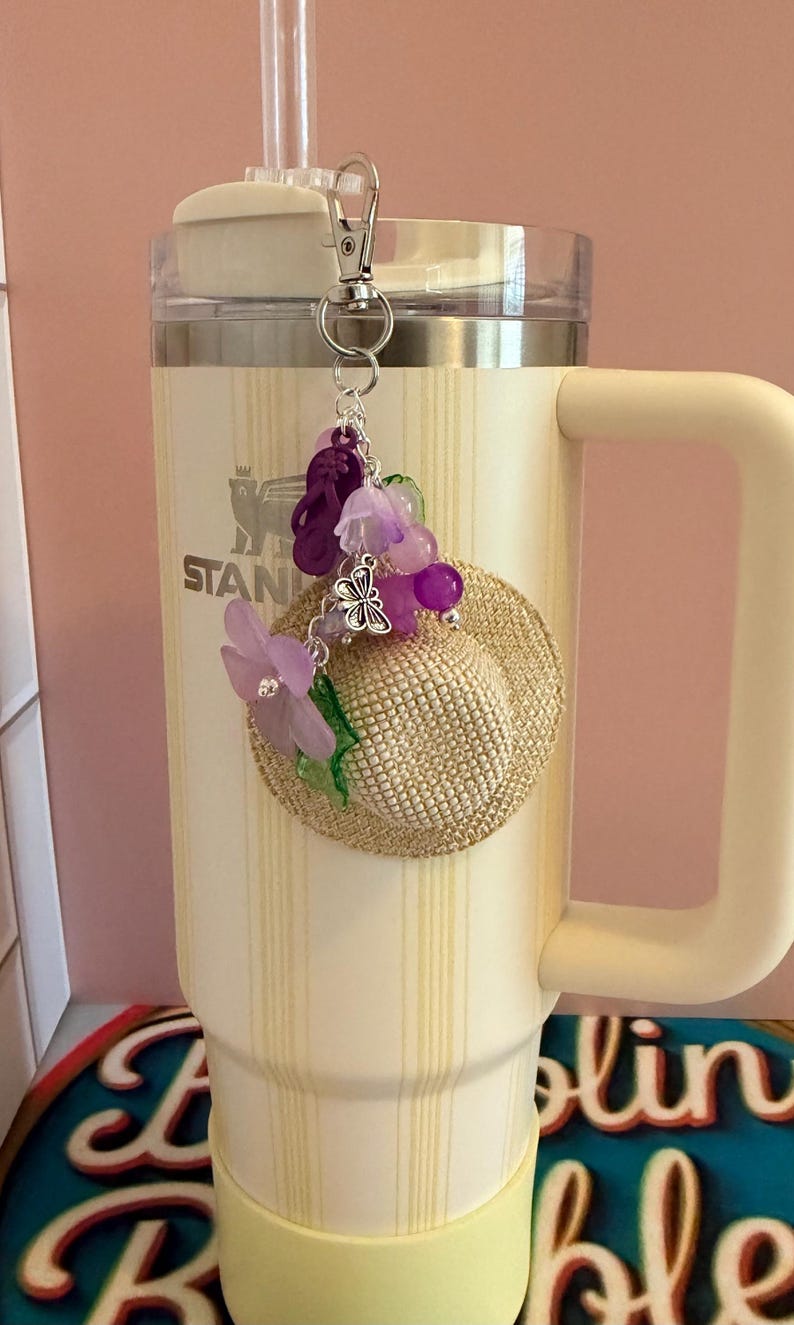 May include: A cream-coloured Stanley tumbler with a straw and handle, decorated with a charm. The charm includes a miniature straw hat, purple flowers, beads, and a butterfly. The tumbler has vertical stripes.