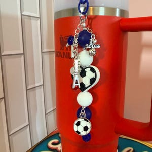 Puede incluir: Un dije para vaso con tem&aacute;tica de f&uacute;tbol que incluye un clip de pl&aacute;stico azul, cadena plateada y cuentas en blanco y azul oscuro. Presenta un dije de coraz&oacute;n, un dije de bal&oacute;n de f&uacute;tbol y un dije plateado con el texto I LOVE SOCCER.