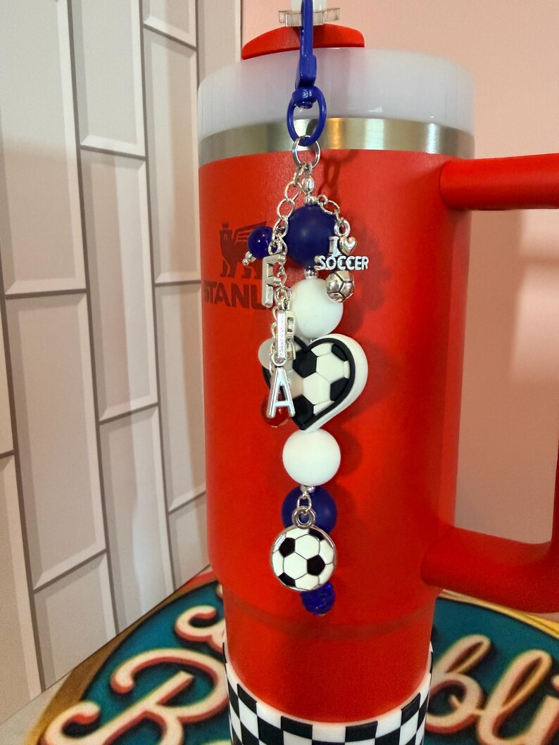 Puede incluir: Un dije para vaso con tem&aacute;tica de f&uacute;tbol que incluye un clip de pl&aacute;stico azul, cadena plateada y cuentas en blanco y azul oscuro. Presenta un dije de coraz&oacute;n, un dije de bal&oacute;n de f&uacute;tbol y un dije plateado con el texto I LOVE SOCCER.