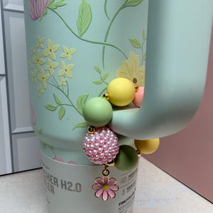May include: A mint green water bottle with a floral design and a beaded charm. The charm features pink pearls, green and yellow beads, and a pink flower pendant. The bottle has the text "CHER H2.0 COOLER".