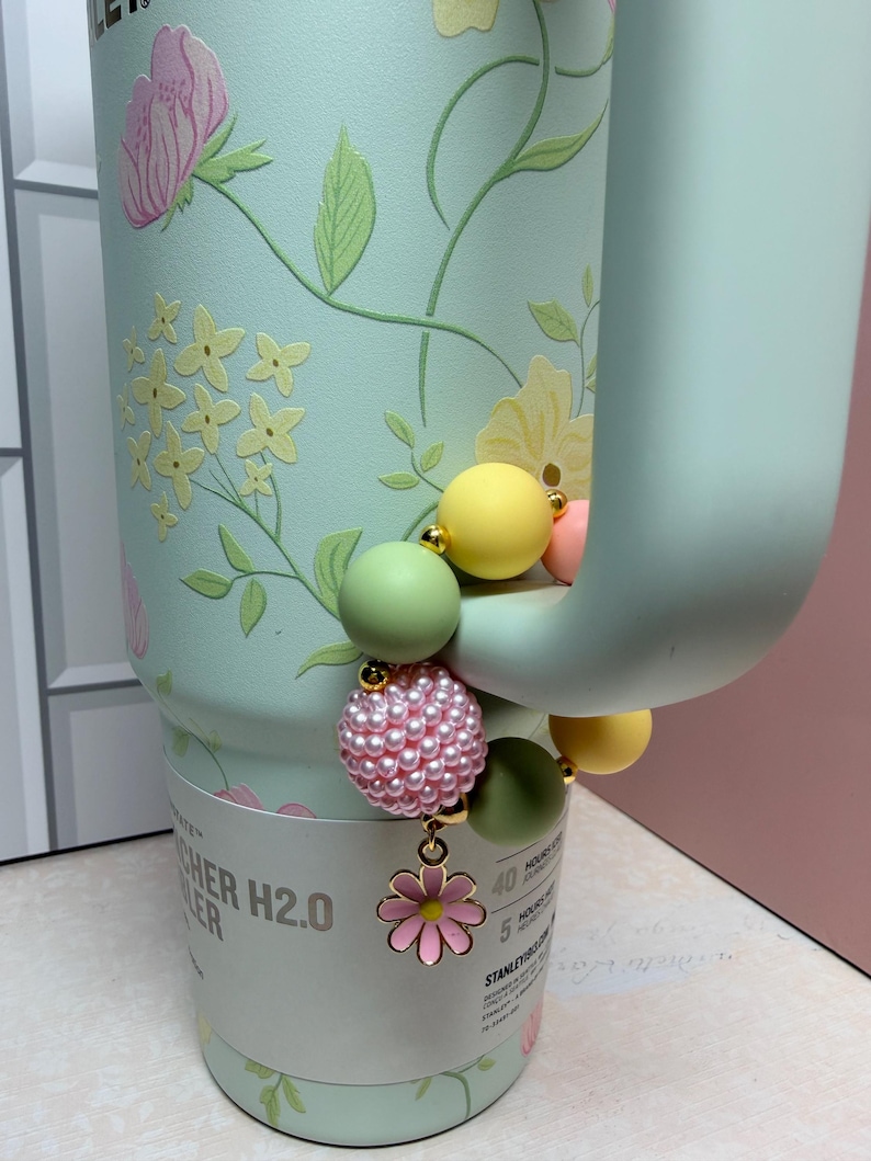 May include: A mint green water bottle with a floral design and a beaded charm. The charm features pink pearls, green and yellow beads, and a pink flower pendant. The bottle has the text "CHER H2.0 COOLER".