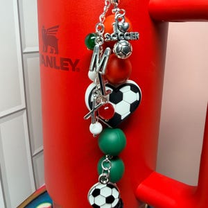 Puede incluir: Un vaso rojo con borde y asa plateados presenta un accesorio colgante. El dije incluye un bal&oacute;n de f&uacute;tbol plateado, un dije de coraz&oacute;n con forma de bal&oacute;n, el texto I LOVE SOCCER, las letras M y X, y varias cuentas de color verde, rojo y blanco.