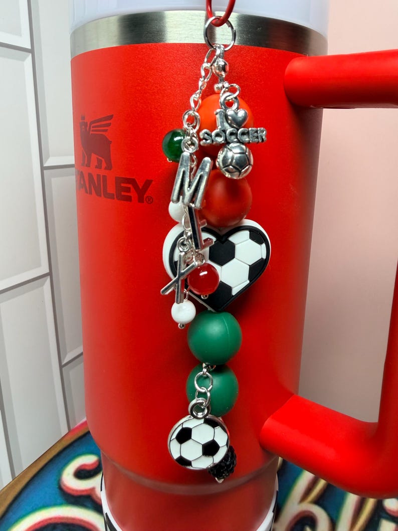Puede incluir: Un vaso rojo con borde y asa plateados presenta un accesorio colgante. El dije incluye un bal&oacute;n de f&uacute;tbol plateado, un dije de coraz&oacute;n con forma de bal&oacute;n, el texto I LOVE SOCCER, las letras M y X, y varias cuentas de color verde, rojo y blanco.