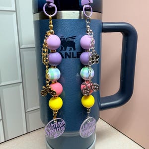 May include: Two beaded cup charms with purple, pink, yellow, and green beads. Each charm has a bicycle charm and a clear oval with the words "Hello Spring" in purple. The charms are attached to a blue insulated tumbler.