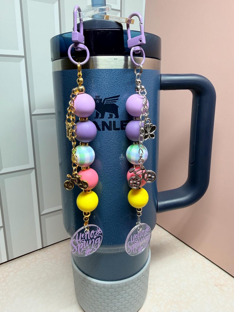 May include: Two beaded cup charms with purple, pink, yellow, and green beads. Each charm has a bicycle charm and a clear oval with the words "Hello Spring" in purple. The charms are attached to a blue insulated tumbler.